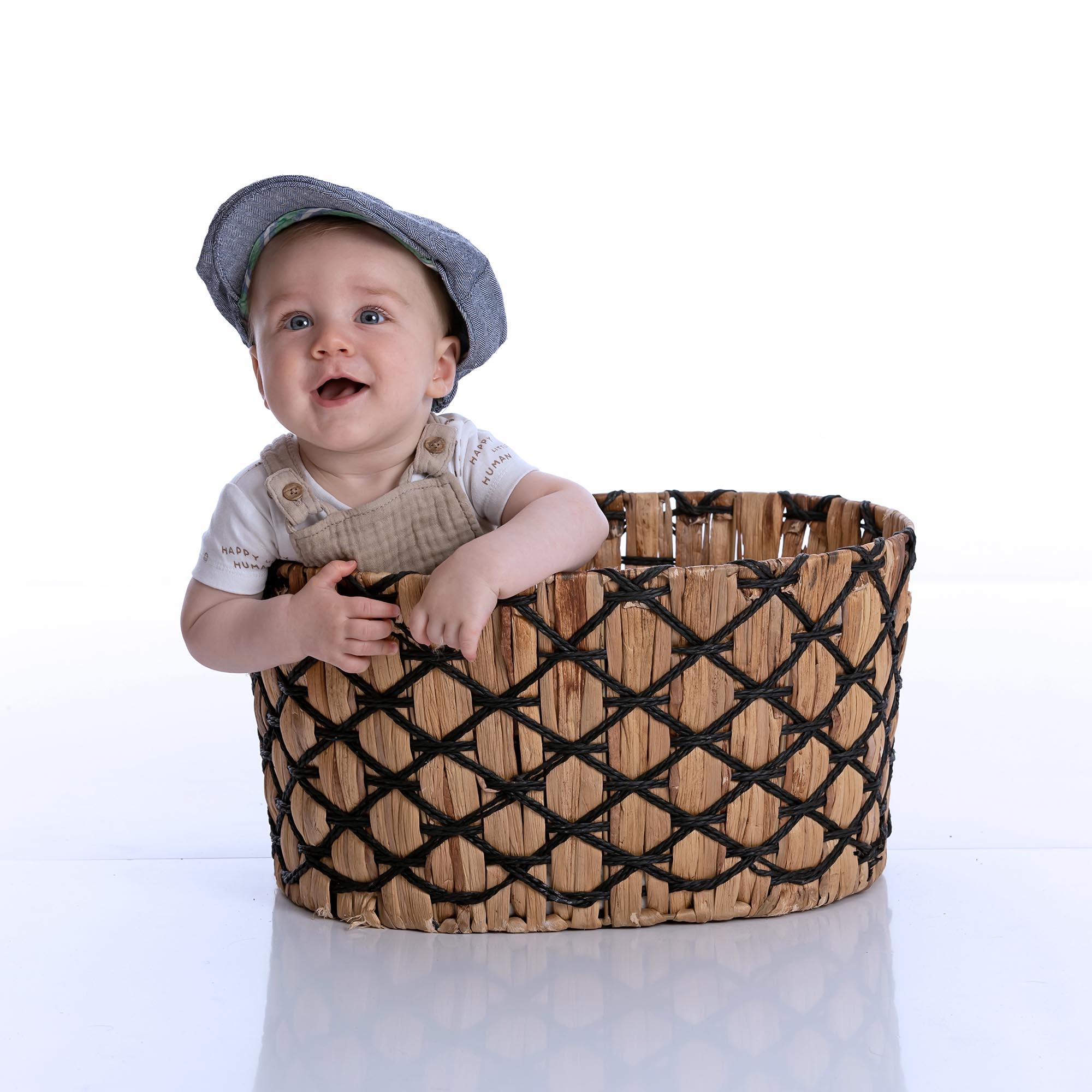 Six month old baby sat in a basket, with a flat cap.